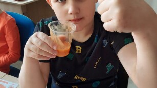Chłopiec trzyma kubek z sokiem marchewkowym i pokazuje gestem, że mu smakuje.