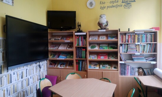 Fotografia biblioteki SPS 26 na ul. Bronowickiej. Widać na niej pomieszczenie zwane czytelnią, wyposażoną w stolik, krzesła oraz półki z książkami. Widać jest również tablicę interaktywną oraz postać mądrej sowy czytającej książkę.